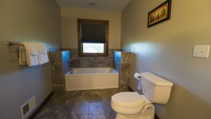 Bathroom with a white tub surrounded by gray stone tiles, a wooden-framed window above, and a towel rack on the left wall.