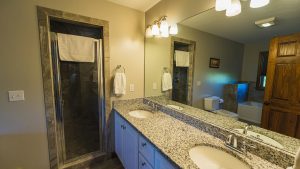 Bathroom with a granite double-sink vanity, large wall mirror, and towel racks; shower stall on the left and toilet on the right; wooden door closed.