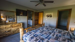 Rustic bedroom with log bed frames, two twin beds, blue plaid quilts, and a wall-mounted TV across from the beds.