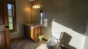 Bathroom with wooden vanity, sink, and mirror; warm light fixture above, window to the yard, and a toilet to the right.