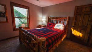 Rustic log bed with plaid red-and-black quilt and bear-themed pillows in a cozy cabin bedroom with a window and lamp.