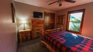 Rustic bedroom with log furniture, a red-and-blue plaid quilt, dresser, wall-mounted TV, and a window overlooking trees.