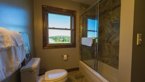 Roomy bathroom with a white toilet, towel on a rack, and a glass‑door shower; window shows a suburban neighborhood outside.