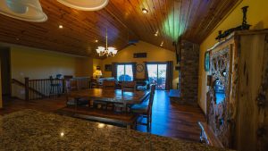 Rustic open-plan living and dining room with a wooden ceiling, chandelier, large dining table, granite island, and a stone fireplace on the right.