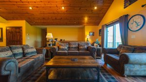 Cozy rustic living room with yellow walls, a wood-paneled ceiling, and leather couches facing a central coffee table. A large window/door opens to a balcony.
