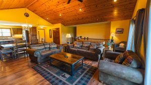 Cozy living room with yellow walls, wooden ceiling, and leather sofas arranged around a coffee table, open to a kitchen and dining area.