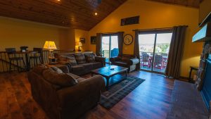 Cozy cabin living room with leather couches, wooden coffee table, and warm lamps, sliding glass doors to a balcony with outdoor seating.