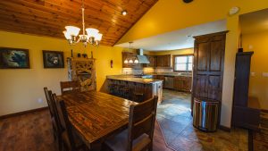 Open-concept kitchen and dining area with a long wooden table, chandelier, and wooden ceiling; yellow walls, granite island, and dark wood cabinets.