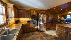 Kitchen featuring dark wood cabinets, granite countertops, and a stainless steel refrigerator with a microwave and oven nearby, illuminated by warm lighting and natural light from a window on the left side.
