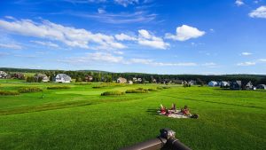 Wide view of a golf course with green fairways, a fire pit with red chairs in the foreground, and houses along the edge under a blue, cloudy sky.
