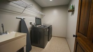 Laundry room with a washing machine and dryer, ironing board, utility sink, and wire shelf along the wall.