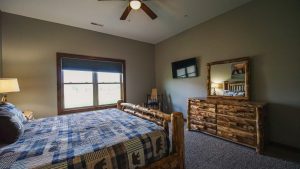 Rustic bedroom with log bed frame and dresser, blue plaid quilt, and a window with wooden trim letting in light.