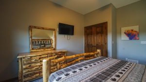 Rustic log-cabin bedroom with a log bed and dresser, a wall-mounted TV, and a colorful dog painting beside wooden double doors on a gray wall.