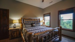 Rustic cabin bedroom with a large log bed, matching nightstands, and warm lamps; forest view visible through the windows.
