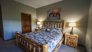 Rustic bedroom with a log bed, blue plaid bear-patterned quilt, and bear-themed pillows; warm lamps on wooden nightstands flank the bed.