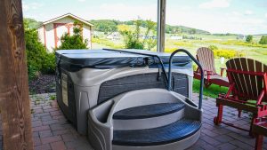 Outdoor hot tub on a brick patio under a wooden shelter, with red Adirondack chairs and a scenic rural landscape beyond.