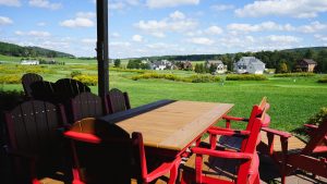 Outdoor patio with red Adirondack chairs around a wooden table, overlooking a green golf course and a row of houses under a blue sky.