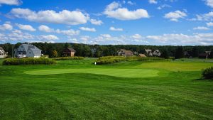 Wide view of a lush golf course with a putting green and flag, houses in the background under a blue sky.