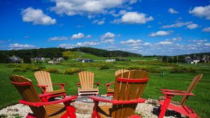 Red and natural wood Adirondack chairs surround a circular fire pit on a green lawn, with a village and rolling hill under a bright, cloudy blue sky.