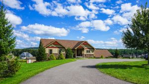 Front view of a beige, ranch-style house with red trim and a curved asphalt driveway under a bright blue sky with white clouds.