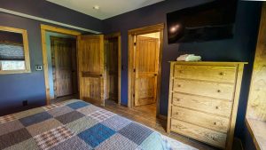 Bedroom with dark blue walls, multiple wooden doors, a tall wooden dresser with towels on top, and a wall-mounted TV above. Patchwork quilt on the bed in the foreground.