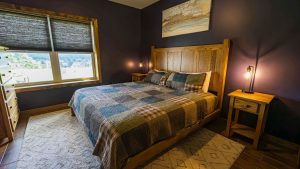 Cozy bedroom with a wooden bed, plaid quilt, and two nightstands on dark purple walls; window with blinds shows an outside neighborhood view.