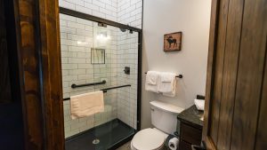 Bathroom with glass shower door, white subway tile walls, and black metal fixtures; white towels on a rail and a rustic moose artwork above the toilet area.