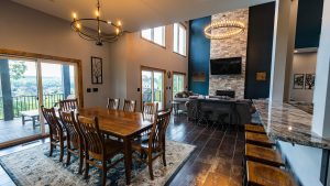 Open-concept dining and living area with a long wooden table, matching chairs, a stone fireplace, and a circular chandelier above.