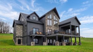 Modern two-story house with dark siding and stone accents on a grassy yard under a blue sky with a large deck and railing