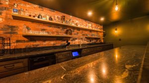 Bar interior with brick wall, liquor bottles on wooden shelves, and glassware under warm lighting by a granite counter.
