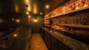Dimly lit bar interior with brick wall, dark marble countertops, bottle shelves, and glassware along the back wall.