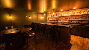 Warm-lit bar interior with brick wall, liquor shelves, and wooden tables and stools.
