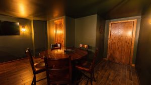 Dimly lit basement dining area with a round wooden table and chairs, a wall-mounted TV, and two wooden doors along olive walls, warm lighting.