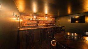 Warm, dimly lit bar interior with a brick back wall, liquor bottles on shelves, and hanging Edison bulbs above a dark wooden counter