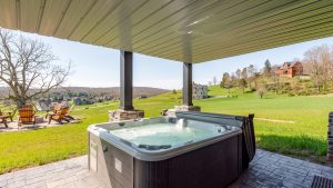 Outdoor hot tub on a stone patio under a sheltered deck, with a green lawn and neighborhood houses in the distance.
