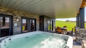 Covered patio with a large hot tub, dining table and chairs along a stone wall, open to green rolling fields.