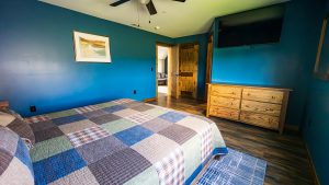 Blue-walled bedroom with a patchwork quilted bed, dresser, ceiling fan, and a wall-mounted TV in view to the right.