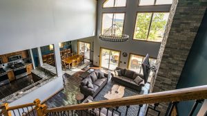 Two‑story open living space with vaulted ceiling, circular chandelier, large windows, and a kitchen island adjacent to a dining area plus a stone accent wall in view.