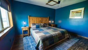 Blue-walled bedroom with a wooden headboard, plaid quilt, and two nightstands with lamps on each side of the bed.