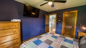 Bedroom with dark blue walls, ceiling fan, and a wall-mounted TV; wooden dresser and doors flank a plaid quilted bedspread in foreground.