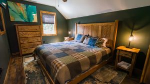 Queen-sized rustic bedroom with plaid quilt, wooden headboard, matching nightstands, and a dresser against dark green walls. A window with a shade and a blue-green abstract painting hang above the dresser, plus warm bedside lamps on each side.