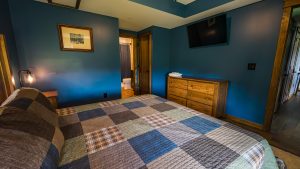 Blue-toned bedroom featuring a patchwork quilted bed, wooden dresser with towels, and a wall-mounted TV; open door reveals a bathroom nearby.