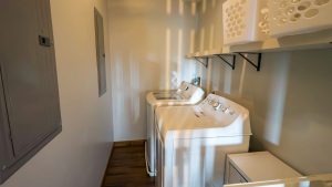 crooked-oak-lodge-044-laundry-room-01 | Swack Vacations Small laundry room with a top-loading washer and front-loading dryer side by side under a shelf with white laundry baskets on the right.