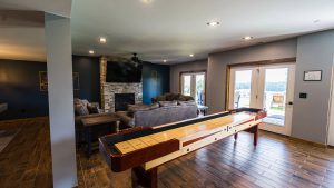 Indoor game room with a long shuffleboard table, leather sofas, and a stone fireplace with a TV above it.
