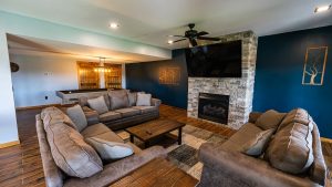 Cozy living room with an L-shaped gray sectional around a wooden coffee table, dark blue accent wall, stone fireplace, and a large wall-mounted TV.