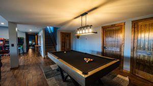 Basement game room with a pool table in foreground, billiard balls racked on the table, and a row of wooden cabinet doors along the wall.