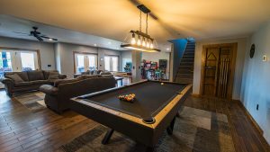 Basement game room with a pool table in the foreground, brown leather sofas, arcade machines in the back, and a staircase along the wall.