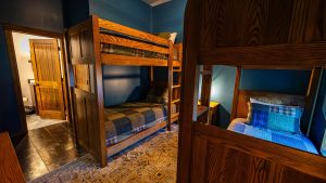 Interior of a blue-walled room with tall wooden bunk beds and plaid bedding; doorway to a lit bathroom visible to the left.
