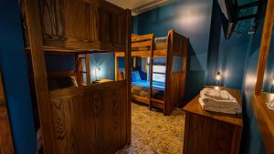 Dorm-style room with blue walls and stacked wooden bunk beds, warm lamps, and folded towels on a dresser/toweling area under soft light.