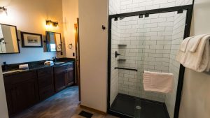 Wide view of a modern bathroom showing a dark wood double vanity with black countertops on the left and a glass-enclosed shower with white subway tiles on the right.